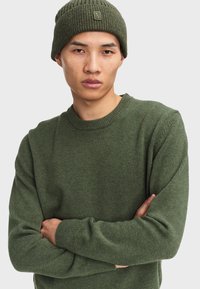 Maglione verde a maglia con scollatura rotonda, polsini a costine e una texture morbida. Indossato con un berretto a coste coordinato con un piccolo logo.