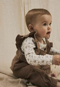 Rjavi kostim iz korduroja z volančki preko cvetne majice z dolgimi rokavi. Mehak material. Dojenček sedi na nevtralni tkanini.
