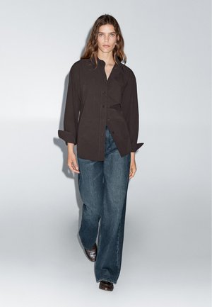 Camicia nera con bottoni, dal taglio unico, abbinata a jeans in denim blu a gamba larga. Il tessuto appare liscio con linee strutturate e polsini.