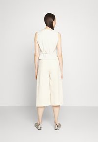 Beige toppi ilman hiuksia, jossa on selässä solmu, yhdistetty leveälahkeisiin beigeihin housuihin ja kuviollisiin slip-on-kengiin, mallinnettuna takaa yksinkertaista taustaa vasten.