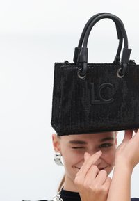 Une personne souriante fait un clin d'œil tout en tenant un sac à main noir étincelant avec les lettres "LC", adoptant une pose enjouée devant un fond blanc.