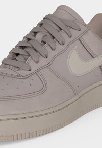 Κοντινό πλάνο μπεζ αθλητικού παπουτσιού Nike με κορδόνια, ραφές, οπές κοντά στη μύτη και λευκό λογότυπο Nike στη πλευρά.