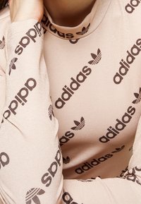 Strakvallend shirt met lange mouwen in lichtroze met glanzende textuur. Voorzien van herhaald donkerbruin "adidas"-tekst en logo, hoogsluitende halslijn.