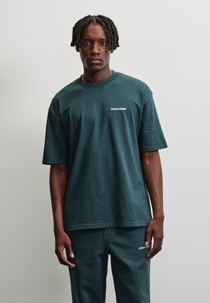 Homme aux cheveux tressés portant un t-shirt vert foncé de la marque Pegador et un pantalon assorti, debout sur un fond clair uni.