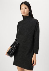 Robe en tricot noir avec col montant et manches longues, ornée de détails en bouton doré sur l'épaule. Fini texturé et coupe fluide.