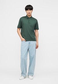 Homme portant un polo vert foncé à manches courtes, un jean large bleu clair et des baskets blanches, debout avec une main dans la poche.
