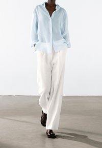 Camicia azzurra in lino con bottoni, pantaloni bianchi larghi, sandali neri. Maniche lunghe e silhouette rilassata, design minimalista.