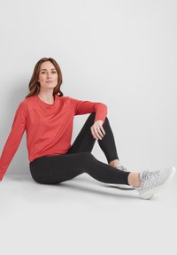 Red long-sleeve shirt with a round neckline, paired with black leggings. The model wears gray athletic shoes with a textured finish.