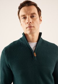 Dunkelgrüner Reißverschluss-Pullover mit texturiertem oberen Abschnitt und glattem unteren Abschnitt. Verfügt über einen kurzen Reißverschluss mit einem Leder-Ziehband.