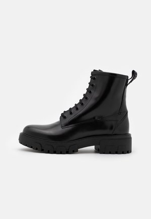 Bota de combate de cuero negro brillante con cordones, suela gruesa con tacos y lengüeta en el talón, vista lateral sobre fondo blanco.