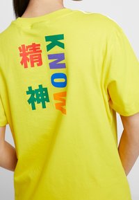 Gul bomulls-T-shirt med färgstark text i olika typsnitt på baksidan: "KNOW" i grönt, lila, orange och rött med kinesiska tecken.