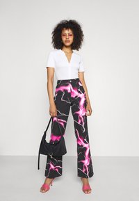 Calças pretas com um padrão de relâmpagos rosa, combinadas com uma camisa branca de mangas curtas e canelada. A modelo segura uma bolsa preta e usa sandálias rosas.