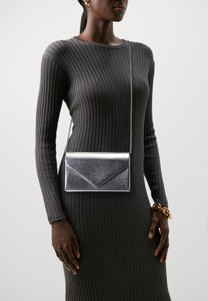 Silberne metallische Umschlagtasche im Envelope-Stil mit dünnem Kettenriemen, ausgestattet mit einer glatten Textur und einem kantigen Klappdetail, getragen zu einem grauen Kleid.