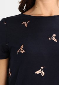 Námořnické tričko s krátkým rukávem a kulatým výstřihem, zdobené vzorem malých, světle růžových kolibříků vyšitých na látce.