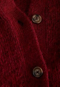 Strickjacke in tiefrot mit einem strukturierten Muster, ausgestattet mit einem dunklen Knopf am Kragen und einem kleinen Knopfloch-Detail.