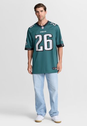 Man staat en draagt een groen Philadelphia Eagles voetbalshirt met nummer 26, lichtblauwe spijkerbroek en witte sneakers tegen een effen achtergrond.