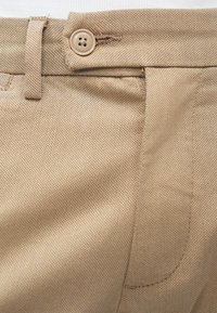 Pantaloni di cotone beige con tessuto testurizzato, dotati di chiusura con bottone e dettagli di cucitura sottili. Vista ravvicinata dell'area della cintura.