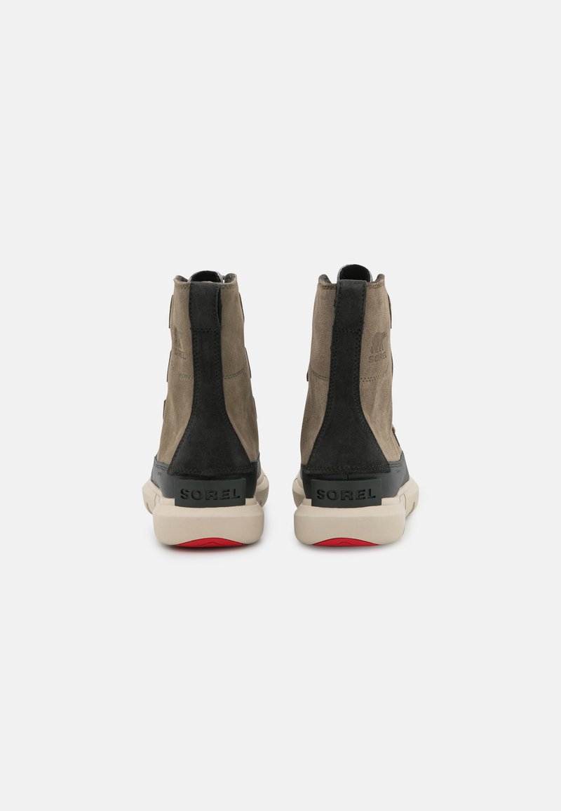 Un paio di stivali in suede neri e beige per l'inverno con suole bianche, visti da dietro su uno sfondo chiaro e semplice.