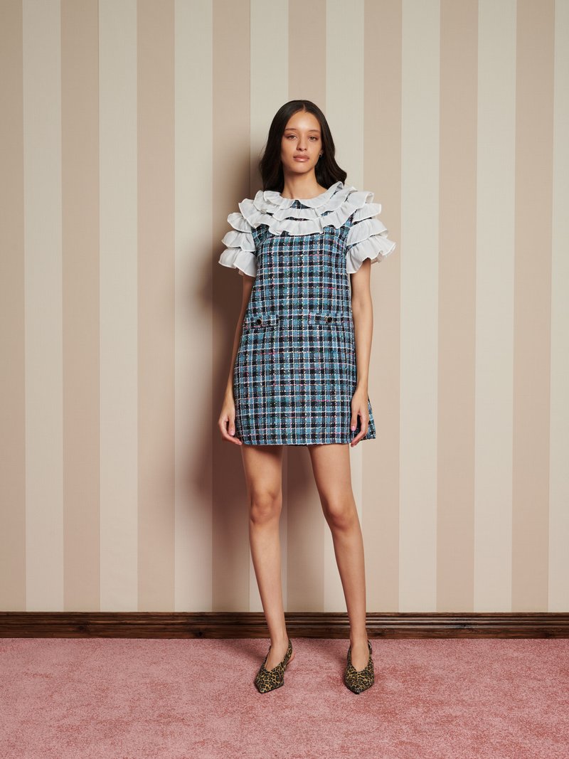 Γυναίκα στέκεται εναντίον ενός μπεζ ριγέ τοίχου, φορώντας μπλε καρό μίνι φόρεμα με στρώματα από λευκές ραμμένες μανίκες και μυτερές γόβες με λεοπάρ σχέδιο.