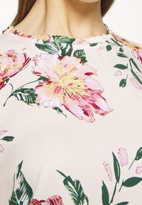 Blusa floreale realizzata in morbido tessuto rosa chiaro con grandi fiori rosa e gialli, foglie verdi e un dettaglio di scollatura arricciata.