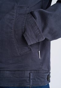 Hand inside the front pocket of a dark blue jacket with visible stitching and a buttoned adjustable hem.