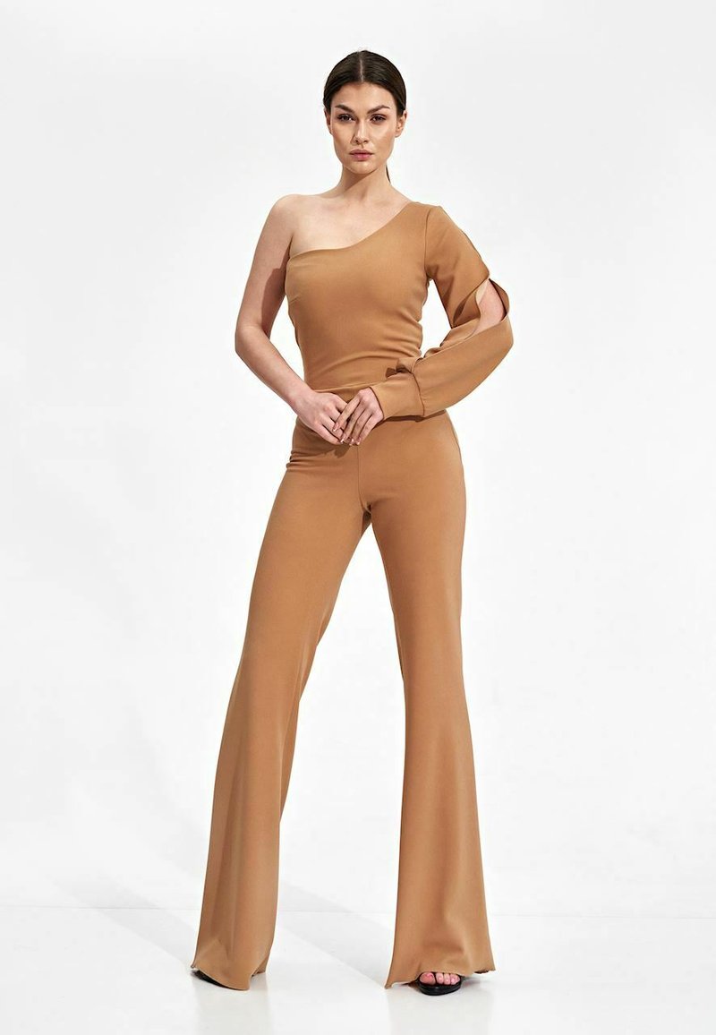 Tan jumpsuit met een asymmetrisch ontwerp met één schouder, lange mouw met uitsnijdingen en flared pijpen, gemaakt van soepel stretchmateriaal.
