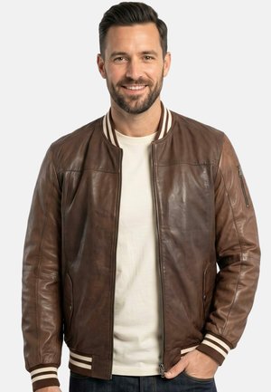 Lächelnder Mann trägt eine braune Leder-Bomberjacke mit gestreiften Bündchen und Kragen über einem weißen Hemd, steht mit einer Hand in der Tasche.