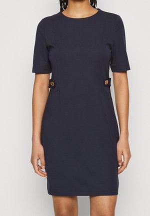 Vrouw draagt een aansluitende marineblauwe jurk met korte mouwen, ronde halslijn en gouden knoopdetails op de taillebandjes, staand tegen een effen achtergrond.