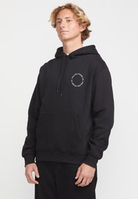 Sweat-shirt à capuche noir avec une poche frontale, un cordon de serrage, et un logo circulaire blanc sur la zone gauche de la poitrine. Fabriqué en tissu doux.