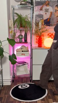 Estante de metal com iluminação rosa, exibindo uma bola de discoteca, lâmpada, plantas e sapatos. Tapete em preto e branco com a forma de uma bola 8 no chão de madeira.