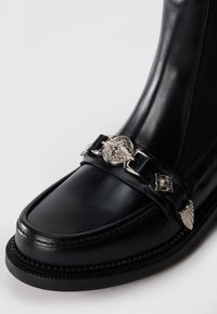 Bottine en cuir noir avec une finition brillante, dotée d'une chaîne décorative en argent et d'accents en emblème à l'avant, ainsi que d'une couture texturée.