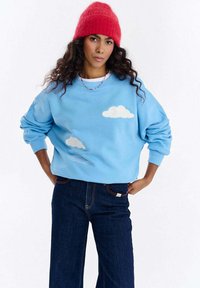 Niebieska bluza z aplikacjami białych chmur, luźny krój, okrągły dekolt, prążkowane mankiety, zestawiona z szerokimi spodniami z ciemnego denimu. Czerwona dziergana czapka beanie.