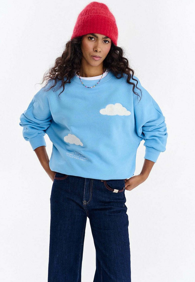 Niebieska bluza z aplikacjami białych chmur, luźny krój, okrągły dekolt, prążkowane mankiety, zestawiona z szerokimi spodniami z ciemnego denimu. Czerwona dziergana czapka beanie.