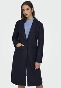 Cappotto di lana navy con taglio lungo, reperto a punte, chiusura a singolo bottone e tasche laterali, abbinato a un maglione texturizzato azzurro chiaro sotto.