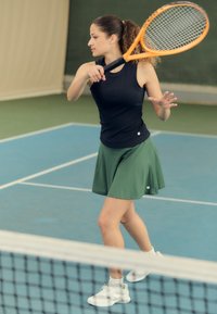 Tennis player in a black sleeveless top and green skirt, holding an orange racket. White shoes and ankle socks. Court background.