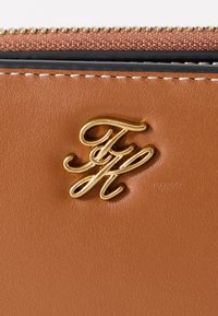 Καφέ δερμάτινο πορτοφόλι με ανάγλυφο χρυσό λογότυπο "FH". Υφή με αντίθετη λεπτομέρεια φερμουάρ στην κορυφή.
