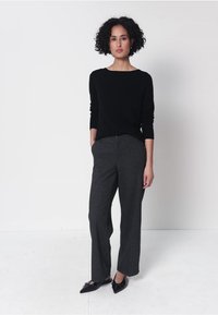 Frau mit lockigem Haar, die ein schwarzes langärmeliges Oberteil, dunkelgraue Hose mit weitem Bein und schwarze spitze flache Schuhe trägt, stehend vor weißem Hintergrund.