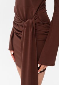 Hnědé přiléhavé šaty s uzlovým detailem v pase, dlouhými rukávy a nařasenou texturou po stranách. Měkká tkanina s hladkým povrchem.