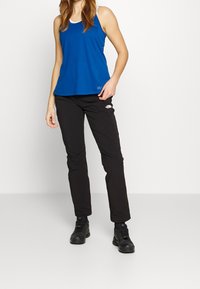 Sininen hihaton paita valkoisilla yksityiskohdilla, yhdistetty mustiin housuihin, joissa on logo. Malli on pukeutunut mustiin ulkoilukenkihin ja seisoo vaalealla taustalla.