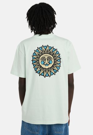 Persona con una camiseta de color claro con un gran gráfico de una cara de sol en azul y beige en la parte trasera, combinada con jeans de mezclilla azul.