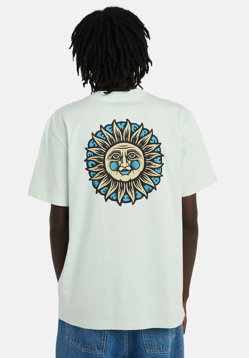 Persona con una camiseta de color claro con un gran gráfico de una cara de sol en azul y beige en la parte trasera, combinada con jeans de mezclilla azul.