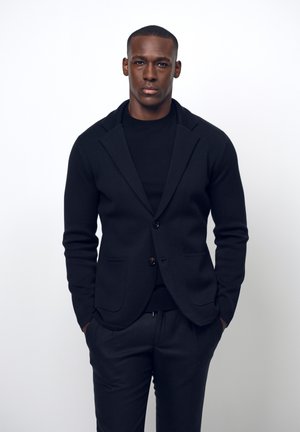 Blazer in pura lana nera con rever a scollo e due tasche anteriori, abbinato a un maglione nero a girocollo e pantaloni, vestibilità slim, design minimalista.