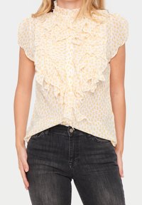 Donna che indossa una camicia senza maniche bianca con pois gialli e volant sul davanti, abbinata a jeans neri scoloriti a vita alta.