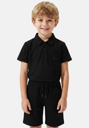 Jeune garçon aux cheveux blonds souriant, portant un polo noir à manches courtes et un short noir à cordon, debout devant un fond blanc.