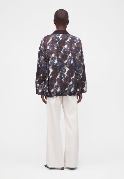 Personne portant une veste ample à motif floral avec des fleurs foncées et bleues et un pantalon blanc à jambes larges, debout de dos.