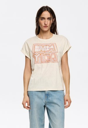 T-shirt beige chiaro a maniche corte, con una grafica testuale colorata "BIMBA Y LOLA" in rosa e arancione, caratterizzata da una texture maculata. Indossato con denim blu.