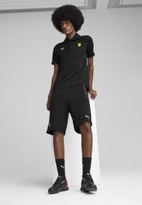 Camiseta polo negra y pantalones cortos, ambos con acentos contrastantes. Logotipos de Ferrari y Puma en la camiseta y los pantalones cortos. Zapatillas deportivas negras con acentos rojos.