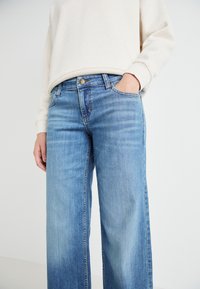 Heledad denim sinine laiemate sääridega teksad, millel on lukuga lend, viis taskut ja õrn tekstuur. Kanda beezhi kapuutsiga pusa.
