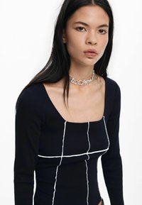 Crna rebrovana majica dugih rukava sa kvadratnim izrezom i bijelim kontrastnim šavovima koji formiraju rešetkastu šaru. Model nosi srebrnu lančićastu ogrlicu.