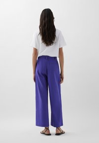 Pantaloni a gamba larga in un vivace tessuto viola, abbinati a una semplice t-shirt bianca. Sandali neri con cinturini completano l'outfit. Texture liscia, stile casual.